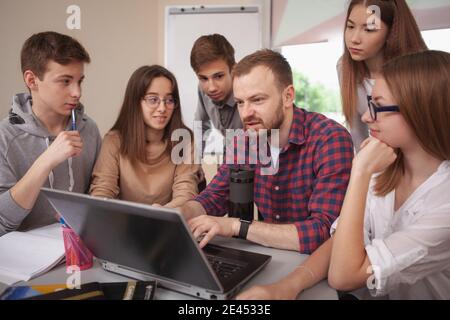 Mature man teaching at high school, using laptop at class, his students gathering around him. Teenage learners enjoying studying with their teacher Stock Photo