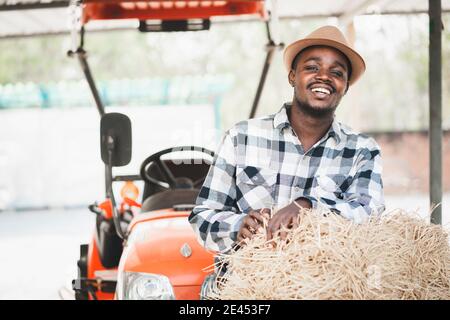 African farmer standing with rice straw bales and tractor. Agriculture or cultivation concept Stock Photo