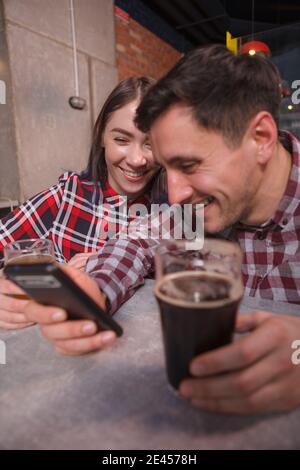 Vertical shot of cheerful young couple laughing, drinking beer and using smart phone Stock Photo