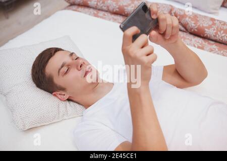 Handsome young man playing online games on his smart phone, lying on a bed Stock Photo