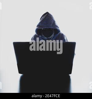 Hacker with a face mask and a hood in front of a laptop - hack attack concept Stock Photo