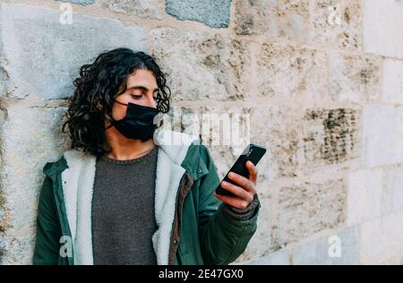 Handsome Long Curly Hair Young Man With Cloth Mask Looking to his Phone Sending Text Messages Leanin Stock Photo