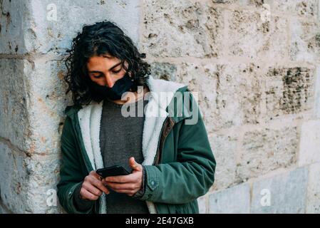 Bored Long Curly Hair Young Man With Cloth Mask Looking to his Phone Sending Text Messages Leaning o Stock Photo