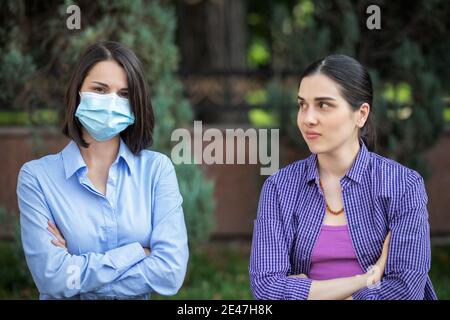 Two young women sits on a summer park bench. One woman wearing a mask, one without a mask. Different attitudes to pandemic. Stock Photo