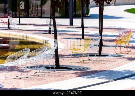 Wicker chairs made of wire stand near a tree on a tile. Nobody is there, unusual chairs are standing in a park. Stock Photo