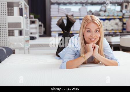 Lovely young woman smiling joyfully, lying on a new bed at furniture store, copy space Stock Photo