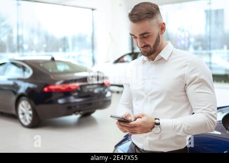 Handsome young man using smart phone at car dealership, copy space. Man texting on his phone, leaning on a car at auto salon Stock Photo
