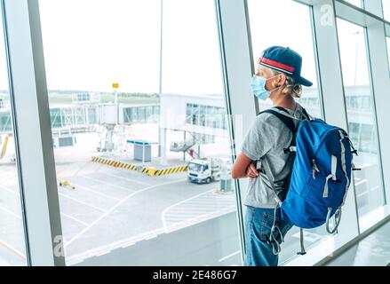 Portrait of lonely teen solo traveler with a backpack in empty airport passenger transfer hall in protective face mask and looking out large windows. Stock Photo