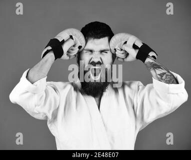 Combat master yells and hits his head. Oriental sports concept. Karate man with mad face in uniform and golden boxing gloves. Man with beard in white kimono on green background. Stock Photo