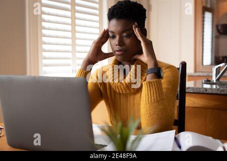 African american woman using laptop rubbing her temples in kitchen Stock Photo