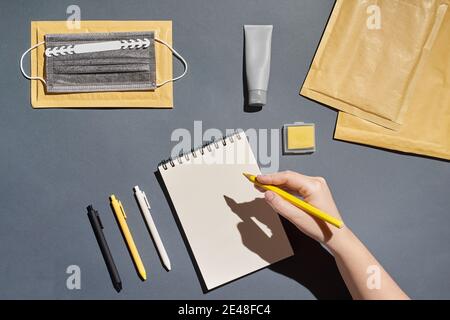 Flat lay notepad, pen, envelope, mask and hand holding pencil on grey background Stock Photo
