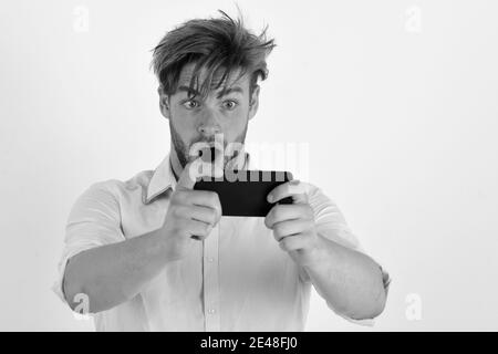 Guy with surprised face holds mobile phone. Young man takes picture with cellphone isolated on white background. Stock Photo