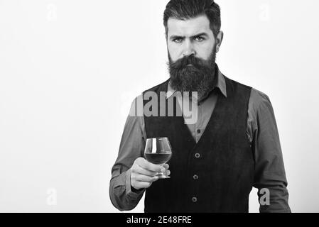 Man with beard holds glass with alcohol on white background, copy space. Alcohol luxury drink concept. Barman in elegant uniform with serious face serves cognac. Waiter with whiskey or scotch in hand. Stock Photo