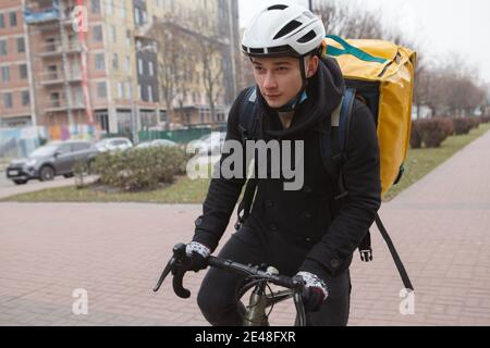 Young deliveryman wearing thermo backpack, cycling city streets in autumn or winter Stock Photo
