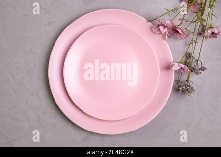 Close-up flat lay of empty pink plate and sprig of pink wildflowers on rough gray concrete or cement background. Spring concept. Selective focus. Stock Photo