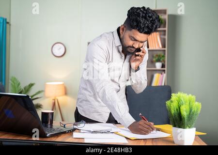 Standing Young man busy making notes while talking on mobile phone in front of working desk - concept of business call, talking with clients noting do Stock Photo