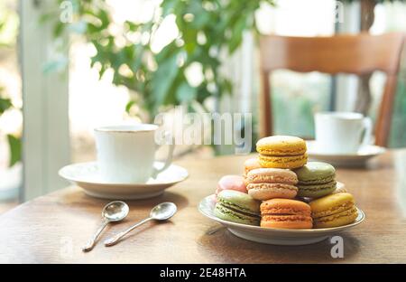 Multicolored macarons or macaroons, traditional French dessert on a plate on a round wooden table with white cups, tasty breakfast Stock Photo