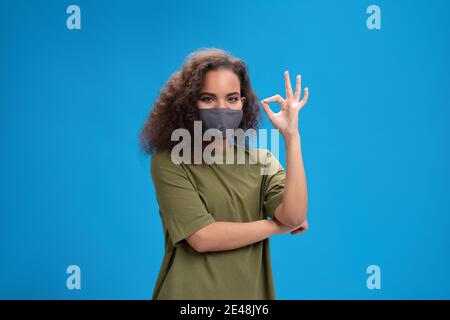 Saying OK using her hands to communicate pretty african american girl in reusable protective face mask. Curly haired young woman showing hello sign Stock Photo