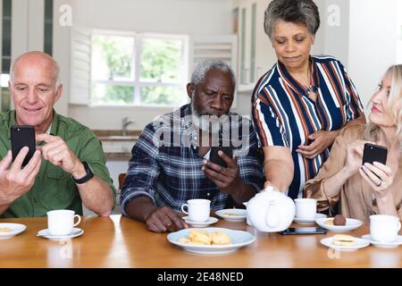 Senior caucasian and african american couples sitting by table drinking tea using smartphone at home Stock Photo