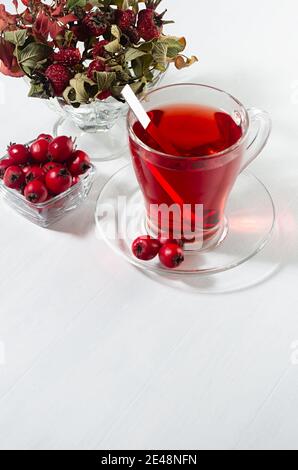 Autumn red herbal tea in transparent mug with hawthorn berries, rose hip, dry leaves in sunlight on white wood board, copy space, vertical, closeup. Stock Photo