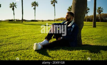 Young guy sitting under tree on green field outdoors Stock Photo