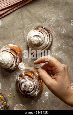 Woman's hand sprinkles icing sugar on fresh baked cinnamon buns. Stock Photo