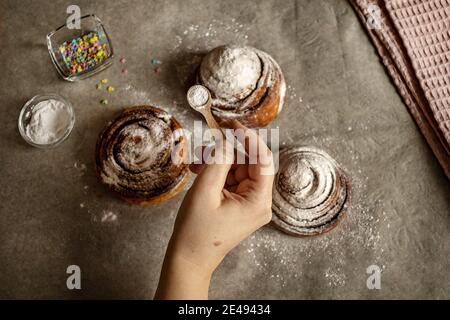Woman's hand sprinkles icing sugar on fresh baked cinnamon buns. Stock Photo
