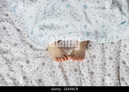 Close-up of toddler girl's feet on the bed under the blanket. Light blue and beige tones Stock Photo