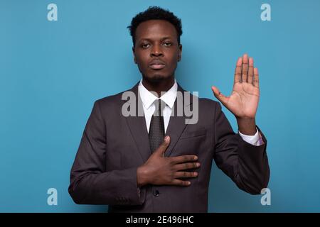 Honest serious man keeps one hand on heart and raises palm, promises or swears to do something. Studio shot on blue wall. Stock Photo