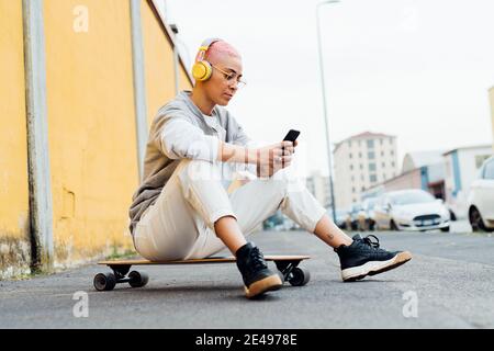 Young woman sitting on skateboard, wearing headphones, using cellphone Stock Photo