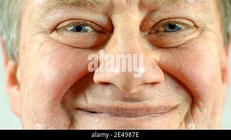 Front portrait of mature adult mans face which smiling.  Wrinkle skin and gray eye, looking at camera. Selective soft focus. Close-up. Indoors. Stock Photo