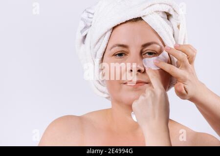 Elderly woman with white towel on her face and hands of beautician applying bandages under her eyes. White background. Stock Photo