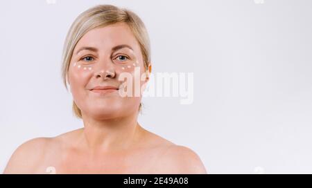 Banner,long format. Portrait of senior blonde woman with drops of moisturizing mask under the eyes against white background with side ad space. Stock Photo