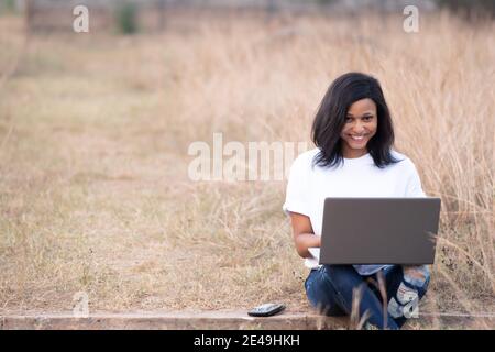 beautiful young african woman, sitting alone outdoor in a park, smiling while using her laptop Stock Photo
