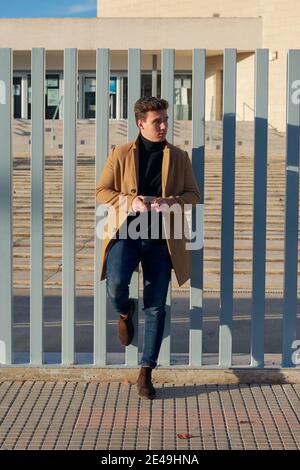 Handsome man dressed in a coat holds his mobile leaning on a fence Stock Photo