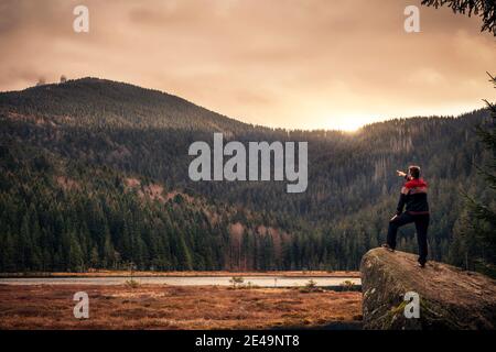 Man Hiker in autumn standing on a rock by the kleiner Arbersee lake and pointing towards peak of großer Arber mountain located at National Park bavarian forest Stock Photo