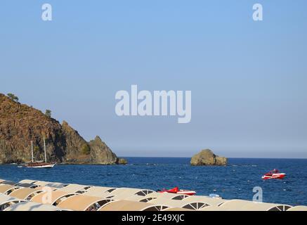 Old wooden sailing ship in sea, blue water. View on mountain and horizon from empty public beach with sunshades. Small jetty boat in Mediterranean Sea Stock Photo