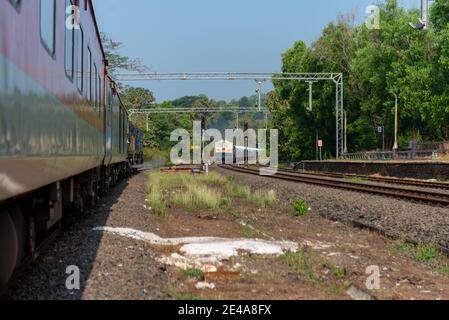 Freight train crossing 02413 Madgaon - Delhi Hazrat Nizamuddin Rajdhani Special at beautiful scenic Pernem Railway Station on Konkan Railway. Stock Photo