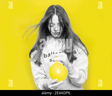 Black and white portrait of young teenage girl with flying hair with yellow balloon blowing air into her face against background in studio. Color Conc Stock Photo