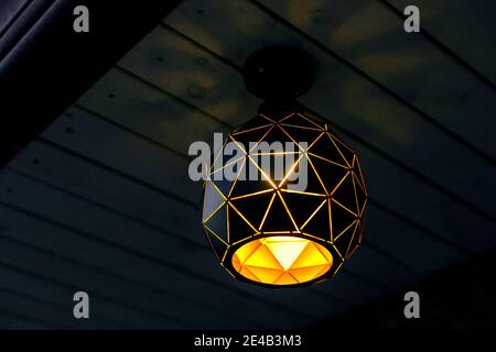 black hanging lantern on wooden ceiling by plank with triangle pattern and warm glow, close-up art of illumination chandelier night scene in backyard Stock Photo