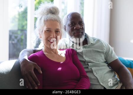 Senior mixed race couple embracing looking at the camera and smiling in living room. staying at home in self isolation during quarantine lockdown. Stock Photo