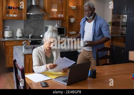 Senior mixed race couple using laptop paying bills together in dining room. staying at home in self isolation during quarantine lockdown. Stock Photo