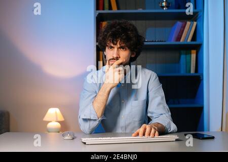 Front view of disgruntled young man typing online message on wireless keyboard, looking at camera. Stock Photo
