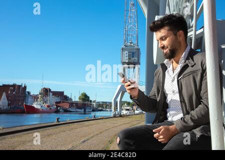 Young man sits alone at the harbor and looks at his smartphone. Stock Photo