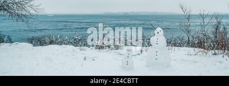 Winter scenery fun outdoor activity for kids, building snowman in snow in Quebec, Canada. Nature fun banner. Two snowmen in front of water Stock Photo