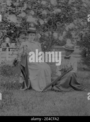 American archive monochrome portrait  of two young women in a garden in front of a blossom tree. One woman is sitting on a chair and one woman is sitting on the grass. The women are holding parasols and wearing skirts and blouses and straw hats. Taken in the late 19th century in Port Byron, NY, USA Stock Photo