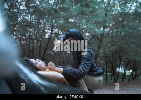a criminals in a mask use a knife coercive and threatening a driver on the hood of a car during a robbery on a lonely road Stock Photo
