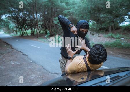 man in a car is robbed by a criminal with a knife while stopping on a lonely road Stock Photo