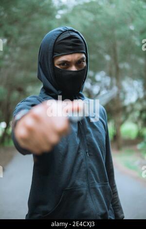 close up of thief in a mask with a knife wearing black hoodie pointed the knife at the camera when stand on the road Stock Photo