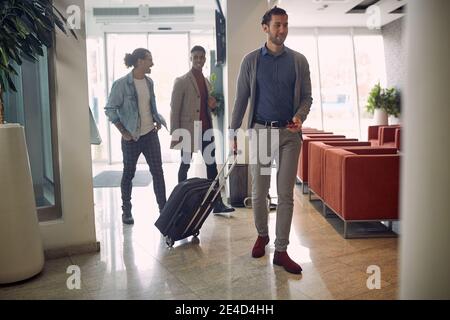 group of young businessmen entering hotel, caring luggage, suitcase on wheels, talking, smiling, laughing